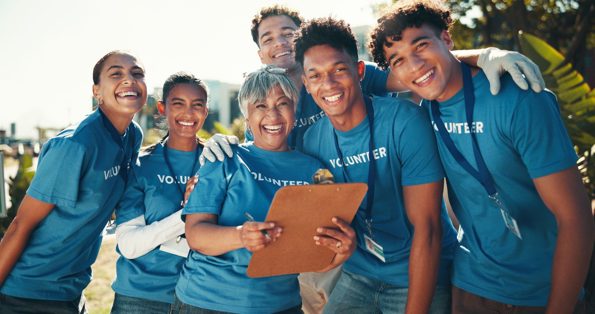 Portrait, happy and group with clipboard, volunteers and joy for collaboration, charity and fundraiser. Outdoor, hug and mature woman with team, smile and good news for community service